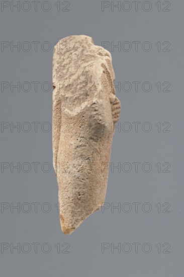 Ushabti - fragment, Mummy-shaped figurine; flat pillar on the back. Preserved central fragment from the shoulder right arm damaged to hip height. Arms crossed on the chest, right folded over the left; only the hands visible from under the shroud. Hoes in both hands with slightly curled ends. In the right hand a smooth rope, from which a trapezoidal basket with a semicircular handle is slung over the left shoulder. Convex tools; engraved basket. Remains of a three-part smooth wig; at the back the wig is slightly wider than the pilaster. Fragmentarily preserved straight, long beard. Engraved inscription placed in a single column on the front of the legs, text bounded by two vertical lines. Tell Atrib Egypt, shabti; height 6 cm, width 3.4 cm., A single elongated, irregularly shaped stone object with a rough, pitted surface is shown against a solid gray background; the object is predominantly beige with areas of light tan and slightly darker brown mottling, and it has a tapered, pointed en...