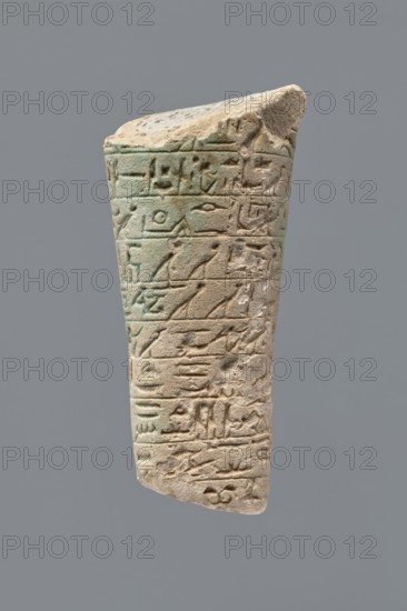 Ushabti - fragment, Fragment of the legs of an ushabti figurine preserved from hips to ankles. The back rests on a pillar. Inscribed hieroglyphs placed in ten horizontal bands around the legs of the figurine, separated by thin lines. Mummy-shaped figurine used as grave goods, with depictions of hoes. Ptolemaic Period, 332–30 BC. Found at Tell Atrib. Ushabti; height 8.5 cm, width 3.8 cm., A worn vertical stone fragment with a tapered shape and a rough broken top edge, showing carved horizontal bands of shallow incisions and raised shapes; the surface displays a mix of light beige, sandy tones and areas of pale green patina, with subtle brownish spots and darker weathered edges; the texture alternates between smoother glazed-looking patches and rougher eroded areas, and the fragment is shown against a uniform grey background.