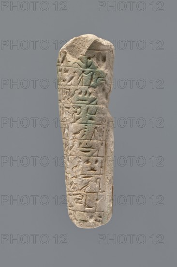 Ushabti - fragment. Mummy-shaped figurine fragment of the legs preserved from hips to ankles; the back rests on a pillar. Inscribed hieroglyphs in ten horizontal bands around the legs, separated by thin lines. Function: ushabti; grave good. Materials: probably faience/stone not specified. Date: 332–30 BC, Ptolemaic Period. Place of discovery: Tell Atrib. Dimensions: height 8.5 cm, width 3.8 cm., A narrow, elongated stone object with a rounded top and bottom is shown against a plain gray background; the stone is light beige to sandy in color with areas of pale greenish discoloration, and its surface features several carved rectangular panels and shallow relief shapes arranged in vertical rows, with some edges chipped and worn, giving an overall weathered, pitted texture.