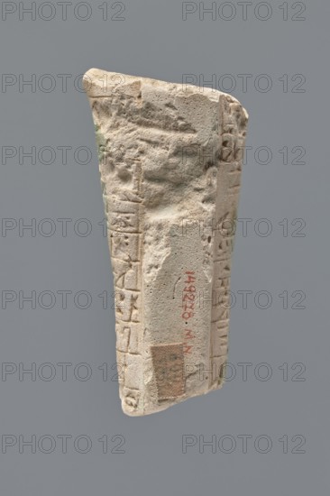 Ushabti - fragment, Fragment of the legs of an ushabti figurine preserved from hips to ankles. The back rests on a pillar. Inscribed hieroglyphs arranged in ten horizontal bands around the legs, separated by thin lines. Mummy-shaped figurine, grave good; hoes. Ptolemaic Period, 332–30 BC. Excavated at Tell Atrib. Ushabti; height 8.5 cm, width 3.8 cm., A roughly rectangular fragment of stone with an uneven, weathered surface and worn edges, showing carved grooves and shallow incised patterns along its sides; the stone is primarily light beige with patches of darker tan and small areas of pale green discoloration; the background is a solid medium gray.