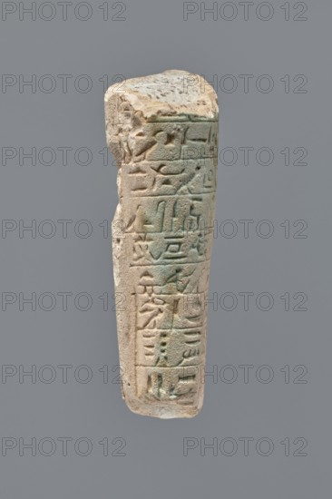 Ushabti - fragment, unknown role classification of creator, Mummy-shaped figurine, grave good. Fragment of the legs of an ushabti preserved from hips to ankles. The back rests on a pillar. Inscribed hieroglyphs placed in ten horizontal bands around the legs, separated by thin lines. 332–30 BC; Ptolemaic Period. Tell Atrib; ushabti. Height 8.5 cm, width 3.8 cm., A weathered vertical stone fragment with carved grooves and shallow reliefs, showing a worn top edge and an irregular side profile; the surface displays a mix of light beige, sandy tan, pale gray, and areas of greenish patina, with subtle darker brown spots and shadows accentuating the carved lines and recessed areas.
