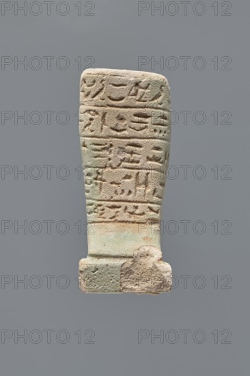 Ushabti - fragment, Mummy-shaped figurine, grave good. Fragment of the legs preserved from the knees down; on the back a pilaster, the feet on a base. Imprinted hieroglyphs in five bands around the legs. 332–30 BC; Ptolemaic Period. Found at Tell Atrib. Ushabti. Height 7.2 cm, width 3 cm., A vertical carved object with a rectangular top tapering slightly toward a wider base, showing a weathered surface with shallow incised marks arranged in horizontal bands; the material appears grainy and worn with areas of rougher texture and small pits, and a chunk missing near the lower right edge; the dominant colors visible are pale beige, light tan, and soft green, set against a plain neutral gray background.