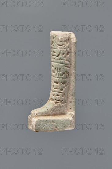 Ushabti - fragment. Mummy-shaped figurine, fragment of the legs preserved from the knees down. On the back a pilaster; feet on a base. Imprinted hieroglyphs in five bands around the legs of the figurine. Ptolemaic Period, 332–30 BC. Tell Atrib. Ushabti. Height 7.2 cm; width 3 cm., A small sculpted fragment of a leg and foot mounted on a rectangular base, shown against a neutral gray background, with the piece appearing in pale beige and light greenish-blue tones; the vertical portion displays shallow carved recesses and raised textured shapes along its front face, while the foot is rounded and rests on a stepped platform that shares the same muted beige and green coloration, with subtle surface pitting and worn edges visible.
