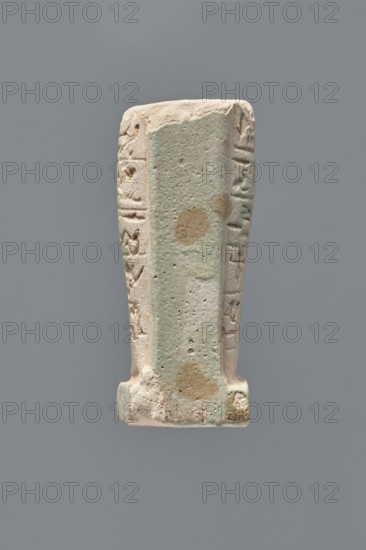 Ushabti - fragment, Mummy-shaped figurine ushabti, fragment of the legs preserved from the knees down. On the back there is a pilaster; the feet rest on a base. Imprinted hieroglyphs in five bands around the legs. Ptolemaic Period, 332–30 BC; Tell Atrib. Height 7.2 cm, width 3 cm., An upright rectangular carved object with a slightly tapered form, showing a textured central surface with a pale greenish tint and worn tan patches, flanked by rougher off-white edges carved with shallow linear impressions and raised decorative elements; the base is slightly wider and shows similar pale green and tan discoloration with rough, chipped areas, all set against a flat neutral gray background.
