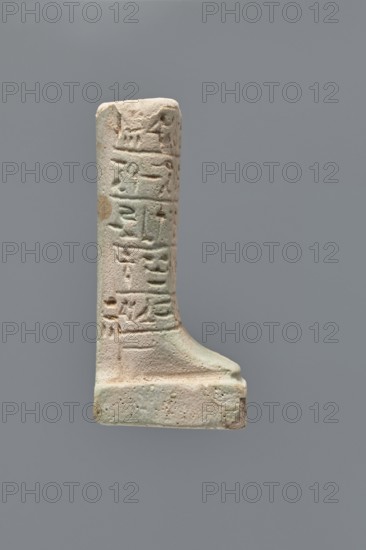 Ushabti fragment, Mummy-shaped figurine grave good. Fragment of the legs preserved from the knees down; on the back a pilaster, feet on a base. Imprinted hieroglyphs in five bands around the legs. Ptolemaic Period, 332–30 BC. Findspot: Tell Atrib. Ushabti. Height 7.2 cm; width 3 cm., A vertical L-shaped stone object photographed against a plain gray background, showing a weathered surface with a few areas of rough texture and pitting; the object is mostly light beige to pale cream with subtle greenish and brownish discolorations in places; one face displays several raised linear and incised shapes forming horizontal bands, and the lower portion broadens into a rectangular base with rounded edges and a gently sloping foot-like projection; the top is flat and slightly irregular, and overall the piece has an aged, worn appearance with small chips and surface variations.