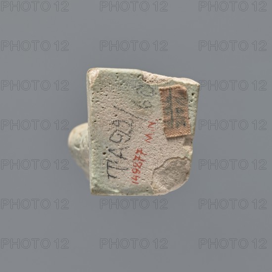 Ushabti fragment, mummy-shaped figurine; fragment of the legs preserved from the knees down. On the back there is a pilaster; the feet on a base. Imprinted hieroglyphs in five bands around the legs. Ushabti used as grave goods. Ptolemaic Period, 332–30 BC. Tell Atrib. Materials/dimensions: height 7.2 cm, width 3 cm., A small worn rectangular object with rounded edges is shown against a gray background; its surface is textured with small pits and irregularities and displays a worn pale greenish-beige color with areas of light beige and off-white; a small rough patch of tan fabric is attached to one corner, and there are smudges and darker gray marks on the surface; the object appears chipped at one edge, revealing a lighter interior, and a faint shadow is cast beneath it.