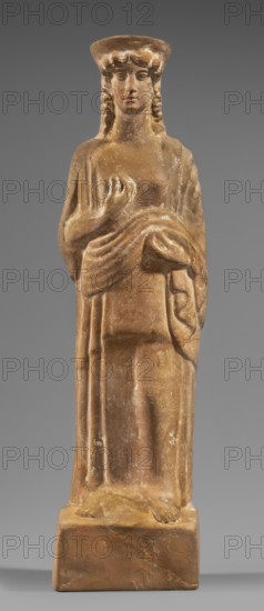 Figurine of a woman with a polos on her head, Tanagra workshop, early 5th century BC circa 480 BC, Tanagra Greece, female figurine, terracotta, height 28.6 cm, width 8 cm, depth 7 cm, weight 0.50 kg., A standing carved figure made of brownish stone with lighter worn areas, depicted wearing a long draped garment and a flat hat, with wavy hair falling to the shoulders; the figure's facial features are visible and neutral in expression, the right hand is raised toward the chest while the left hand holds drapery, and the figure stands barefoot on a rectangular base against a plain gray background.