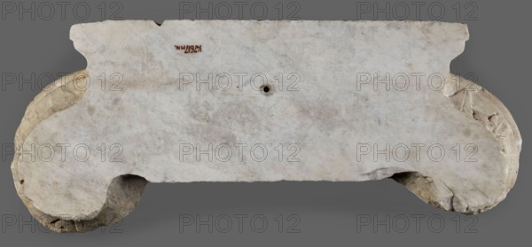 Ionic pilaster capital, unknown Roman workshop role classification of the maker, workshop author; capitals architect, Roman architecture, cymatium ornament, rosettes architect, rosettes ornament, ovoid ornament; 1st century, Roman Empire 27 BC–476; architectural element; element of architectural decoration; capital; height 29.1 cm, width 70 cm, depth 13 cm, weight 26 kg, A weathered stone slab with an irregular rectangular central section and curved ends, shown against a neutral gray background; the slab surface is mostly light gray with patches of off-white, beige, and subtle brown discoloration, showing a rough, pitted texture and a small round hole near the top center; the curved end areas reveal exposed inner stone that is a warmer beige and light brown, with some darker brown crevices and chipped edges.