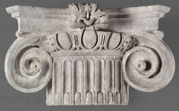 Ionic pilaster capital, unknown Roman workshop, 2nd century, Roman Empire 27 BC–476, architectural element; height 27 cm, width 46.5 cm, depth 18 cm, weight 19.5 kg, height 26 cm, A close-up view of a carved architectural element in shades of gray and off-white, showing a symmetrical design with a central fluted column section and a pair of large volute spirals on either side; a decorative band with rounded and pointed leaf-like motifs sits above the flutes, and a stylized floral or leaf carving is visible at the top center; the surface shows subtle variations in light and shadow, with worn and textured stone appearance and faint darker gray areas in crevices.