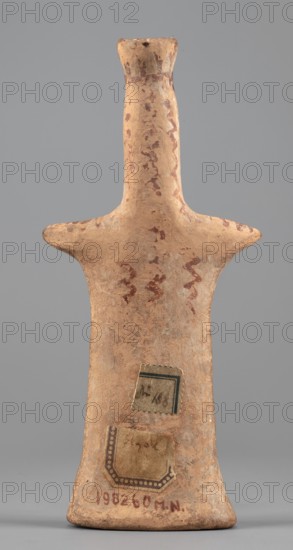 Female idol, Female idol with arms marked as appendages. On a long, cylindrical neck is a nose and an upward-curved strand of hair with a diadem. The robe, breasts, and hair are rendered with a varnish line. Terracotta figurine, 6th century BC previously dated 7th century BC, from Boeotia. Height 16.7 cm; width 7.6 cm; depth 3 cm; weight 0.15 kg., A tall, narrow clay figurine with an elongated neck and a flared top, showing a pair of horizontal protrusions at shoulder level like short arms; the surface is rough and weathered with a base that flares outward. The object displays several irregular reddish-brown painted wavy motifs and spots on a light tan to sandy beige background, with areas of darker brown discoloration and pale patches. A few small rectangular paper labels are affixed near the lower body, showing faded tones of beige and muted green with decorative borders, and there is reddish-brown pigmentation near the base. The background behind the object is a uniform medium gray.
