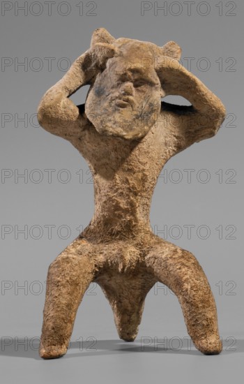 Satyr figurine. Clay figurine depicting a satyr with a face stylized as a mask with protruding ears and two horns. The man is shown crouching with legs widely apart, supported by a thick tail. The hands are raised upwards, emphasizing and exposing the figure's genitals; the satyr's hands touch the head, which is tilted slightly to the left. The face has lush, regular facial hair; small eyes; clearly defined eyebrows and broad nose; a thin closed mouth. Hands and legs are treated schematically compared with the carefully modeled face. Made of fine-grained yellowish-tinted clay. 5th century BC previous datings: 2nd half of the 6th century BC–beginning of the 5th century BC; earlier: end of the 6th century BC–first half of the 5th century BC. Place of origin: Corinth. Figurine. Height 8.8 cm; width 4.8 cm; depth 4.4 cm; weight 0.04 kg., A weathered sculptural figure carved from a rough tan and brown material stands on a flat gray surface against a neutral gray background; the figure has a...