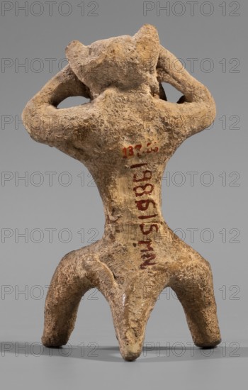 Satyr figurine, clay; depicts a satyr with a face stylized as a mask with protruding ears and two horns. The crouching male figure has legs widely apart, supported by a thick tail; hands are raised and touch the head tilted slightly left, emphasizing and exposing the genitals. Face with lush, regular facial hair; small eyes, clearly defined eyebrows and broad nose, thin closed mouth. Hands and legs are treated schematically compared to the carefully modeled face. Made of fine-grained yellowish-tinted clay. Date: 5th century BC previous datings: 2nd half of the 6th century BC–beginning of the 5th century BC; earlier: end of the 6th–first half of the 5th century BC. Place of origin: Corinth. Figurine; height 8.8 cm, width 4.8 cm, depth 4.4 cm; weight 0.04 kg., A small rough-textured figurine with a weathered surface in shades of beige, tan, and brown, shown from the back; the figure has a broad upper body with two raised arms bent so hands rest near the head, a narrow waist, and a lower ...