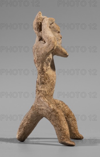 Satyr figurine. Clay figurine depicting a satyr with a face stylized as a mask with protruding ears and two horns. The figure crouches with legs widely apart, supported by a thick tail; hands are raised upwards, touching the head tilted slightly to the left and emphasizing the exposed genitals. Face has lush, regular facial hair; small eyes; clearly defined eyebrows and broad nose; thin closed mouth. Hands and legs are treated schematically compared with the carefully modeled face. Fine-grained yellowish clay. 5th century BC previously dated to late 6th–early 5th century BC. Origin: Corinth. Medium: clay figurine. Dimensions: height 8.8 cm; width 4.8 cm; depth 4.4 cm; weight 0.04 kg., A weathered tan and brown clay figure standing on a flat gray surface against a smooth gray background, with a rough textured surface and uneven coloration; the figure has a vertical torso, elongated bent legs forming a wide stance, and raised arms positioned near its head, with a protruding facial profil...