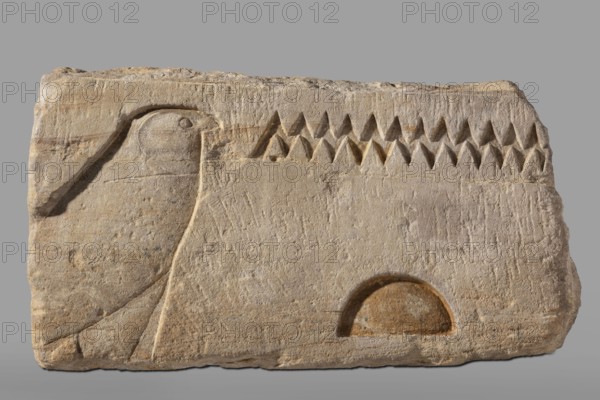 Fragment of an architrave with hieroglyphs from the temple of Horus in Buhen. Horus mythology, falcon iconography, hieroglyphs, Egyptian sacred architecture. Fragment of an architrave with preserved hieroglyphs in recessed relief. From the left, the sign hor, followed by the signs n and t. Traces of yellow polychrome are preserved on the surface. c. 1479–c. 1425 BC; New Kingdom, 18th Dynasty possibly Thutmose III. Findspot: Buhen, Faras site/country of find. Bas-relief, architectural element. Height 31 cm; width 60 cm; depth 52 cm; weight 143.5 kg., A rectangular stone slab in beige and light brown tones with rough, weathered texture shows a low-relief carving of a stylized bird profile on the left, with a rounded head, beak, and a simple carved eye, and a gently curved body and wing lines; across the upper center is a row of carved triangular shapes creating a sawtooth pattern in darker brown shadows; near the lower right there is a semicircular recessed area with warmer brown shading...