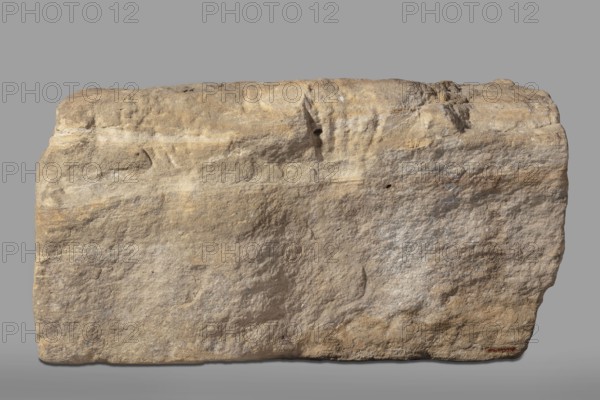 Fragment of an architrave with hieroglyphs from the temple of Horus in Buhen. Horus mythology, falcon iconography, hieroglyphs, Egyptian sacred architecture. Fragment of an architrave with preserved hieroglyphs in recessed relief. From the left, the sign hor, followed by the signs n and t. Traces of yellow polychrome are preserved on the surface. c. 1479–c. 1425 BC; New Kingdom, 18th Dynasty possibly Thutmose III. Findspot: Buhen, bas-relief, architectural element; height 31 cm, width 60 cm, depth 52 cm; weight 143.5 kg., A large rectangular block of rough textured stone fills the frame, with an uneven top edge and slightly rounded corners; the surface shows weathered grooves, pits, and layered striations. The stone displays a range of earthy colors including beige, tan, light brown, sandy yellow, and areas of pale gray and off-white. The background is a flat medium gray, and there is a faint shadow beneath the block.