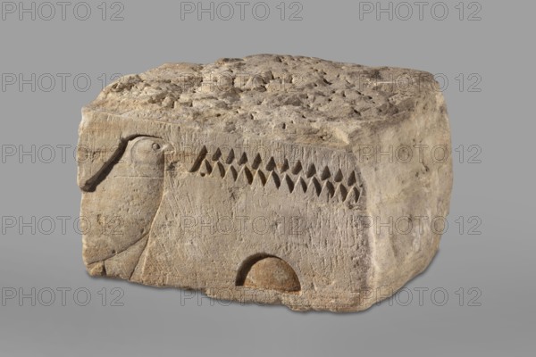 Fragment of an architrave with hieroglyphs from the temple of Horus in Buhen. Horus mythology, falcon iconography, hieroglyphs, Egyptian sacred architecture. Bas-relief architectural element with preserved hieroglyphs in recessed relief: from the left, the sign hor, followed by the signs n and t. Traces of yellow polychrome are preserved on the surface. Circa 1479–1425 BC, New Kingdom, 18th Dynasty possibly Thutmose III. Findspot: Buhen, Faras site type/country of find. Height 31 cm, width 60 cm, depth 52 cm, weight 143.5 kg., A roughly rectangular block of weathered stone in tan and beige tones sits against a neutral gray background, showing a carved relief on one face with the profile of a bird outlined in shallow incisions, a row of repeated triangular indentations, and a semicircular recessed shape near the base; the top surface is uneven and pitted with rough texture, while the sides display smoother areas and worn edges in similar sandy colors.