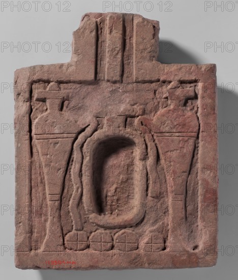 Offering table, Square offering table with a protruding spout. In the center is a cartouche-shaped recess, while on its sides are two relief hes vases with spouts from which water pours. Below are four round loaves of bread. Cartouches. 300 BC–350 AD, Meroitic Period. Faras. Height 9.2 cm; width 32.28 cm., A rectangular carved stone plaque with an irregular top edge showing weathered, rough texture and visible tool marks; the stone is mainly reddish-brown with areas of darker brown and faint gray. The carving includes a central oval recessed cavity framed by raised, rounded borders, flanked by two symmetrical vase-like or tapered vertical forms on either side, each with horizontal incised bands and flat tops. Below the central cavity is a row of rounded, petal-like motifs contained within circular outlines, and vertical fluted or ribbed elements appear above the plaque near the top center. The overall surface shows chips, worn edges, and subtle variations of reddish-brown, brown, and g...