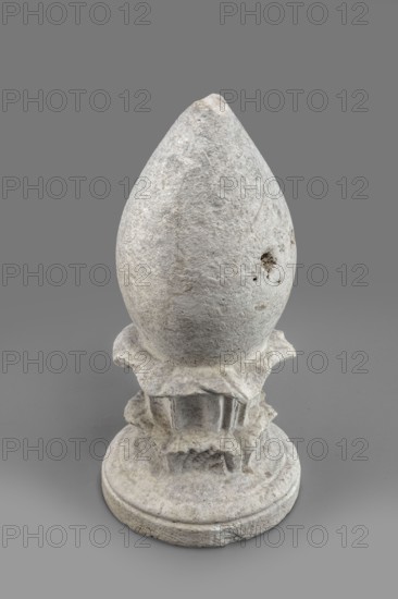 Pine cone-shaped cippus, cippus used in sepulchral funerary architecture, classified as Gallo-Classical scenography/architectural element, Etruria Italy, Praeneste Italy as place of origin, echinus-shaped base from which a double acanthus wreath topped with an egg-shaped element grows, acanthus ornament, 4th–3rd century BC, height 32.3 cm, width 15.2 cm, weight 6.9 kg., A weathered sculpted object with a rough textured surface standing on a round base, shaped like an elongated egg or teardrop atop a decorative, carved pedestal with layered, irregular edges and sculpted ridges; the material appears stone-like with an uneven, pitted surface and small holes on one side. The visible colors are shades of gray and off-white against a mid-gray background.
