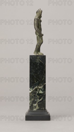Figurine of Poseidon, unknown Roman workshop. Figurine of Poseidon standing in contrapposto, resting on a trident. The weight of the body rests on the left leg; the right is slightly moved back, the foot bent at the toes. The body is nude, correctly modeled, with clearly defined abdominal muscles. In the left hand the figure holds a trident; the right hand is lowered at the side of the body, holding a cylindrical object at hip level. The head is slightly bowed. The face is framed by long hair and a beard. 1st century, Roman Empire 27 BC–476. Terracotta figurine, height 8.7 cm, width 3.5 cm, depth 1.8 cm, weight 0.362 kg with base., A small greenish bronze figure stands in profile atop a tall dark green and black marble plinth with lighter green and white veining, the figure showing a textured mane of hair and a slender body with one arm bent and the other hanging, the plinth resting on a stepped black base against a light gray background.