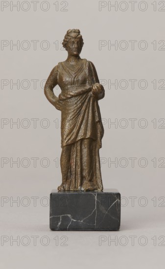 Venus figurine, unknown Roman workshop, Roman period, Roman Empire 27 BC–476, figurine, height 7.4 cm, width 2.8 cm, depth 2.5 cm, A small bronze-colored statue of a standing female figure wearing a draped garment, shown frontally on a dark gray marble base with white veins; the figure has one arm bent with a hand on her hip and the other arm holding a round object near her waist, with visible textured surfaces and folds in the clothing, and the background is a smooth light beige-gray.