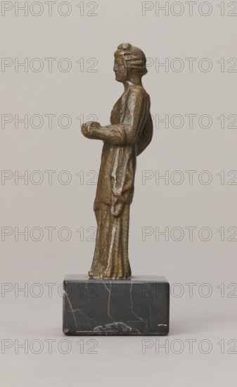 Venus figurine, unknown Roman workshop, Roman period, Roman Empire 27 BC–476, figurine, height 7.4 cm, width 2.8 cm, depth 2.5 cm, A small bronze-toned figurine of a person standing upright on a dark gray to black rectangular stone base; the figure is shown in profile facing left, wearing a long draped garment with visible folds and a textured surface, with hair styled close to the head; the figure’s arms are bent at the elbows, one hand held slightly forward and the other resting near the hip; the base has lighter colored veins and a polished appearance; background is a smooth light beige-gray.