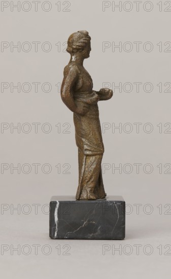 Venus figurine, unknown Roman workshop, Roman period, Roman Empire 27 BC–476, figurine, height 7.4 cm, width 2.8 cm, depth 2.5 cm, A small bronze-colored statuette of a standing person is shown in profile on a dark gray and black marble rectangular base; the figure wears a long dress with visible textured folds and has a relaxed posture with one arm bent and hand resting near the waist and the other arm extended slightly forward holding a rounded object, and the background is a smooth pale beige.