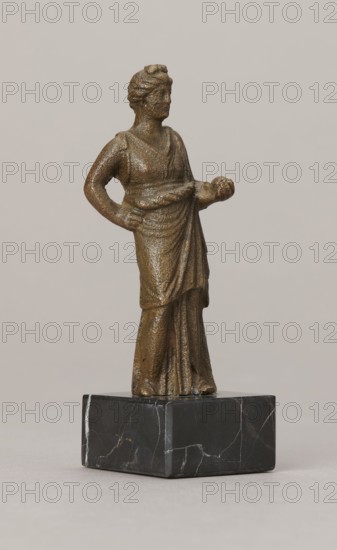 Venus figurine, unknown Roman workshop, Roman period, Roman Empire 27 BC–476, figurine; height 7.4 cm, width 2.8 cm, depth 2.5 cm., A small bronze-colored statuette of a woman wearing a draped gown stands on a dark marble-like rectangular base; the figure has a textured surface, hair styled up, one arm bent with a hand resting on her hip and the other arm extended holding a rounded object, and the overall color palette includes bronze tones, dark gray and black with lighter gray veining on the base, set against a neutral light beige background.