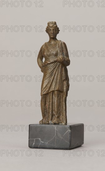 Venus figurine, unknown Roman workshop, Roman period, Roman Empire 27 BC–476, figurine; height 7.4 cm, width 2.8 cm, depth 2.5 cm., A small bronze-colored statuette of a robed female figure stands on a dark gray marble base; the figure wears a long draped garment with visible folds and a sash at the waist, one arm bent with a hand resting on the hip and the other arm extended slightly forward holding a small rounded object, the surface of the statuette shows a textured metallic brown and greenish patina, and the marble base is smooth with lighter gray veining against a neutral pale beige background.