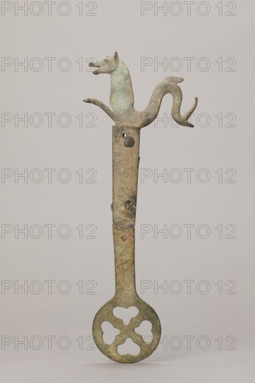 Fragment of a grave brazier, 2nd half of the 5th century, Etruria; height 30.5 cm, width 11.5 cm, depth 2 cm, A vertical metal object with a greenish-brown patina stands against a neutral light gray background; its top ends in a stylized animal head and curved tail forming a symmetrical decorative element, the midsection is a narrow rectangular shaft with visible surface irregularities and a small round protrusion, and the lower end widens into a circular disc pierced with a four-lobed floral-like pattern.