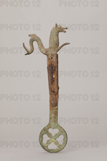 Fragment of a grave brasier, 2nd half of the 5th century, Etruria; height 30.5 cm, width 11.5 cm, depth 2 cm., An upright metal object photographed against a plain light gray background, consisting of a vertical shaft with a decorative animal-shaped top and a rounded perforated base; the top is formed like a stylized creature with a head, open mouth, ears, and a sinuous body that branches into curved limbs, showing a mottled greenish patina; the shaft below the animal has areas of brown rust and rough texture with irregular patches and a few darker corroded sections; the lower portion expands into a circular flat element with four rounded lobed openings, also displaying a greenish surface with spots of brown and darker discoloration.