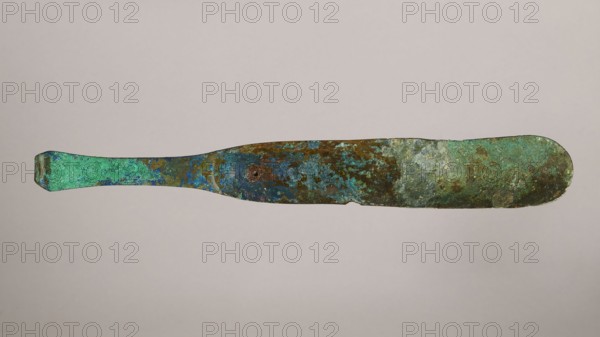 Scraper, metal scraper with a typical slightly curved shape consisting of a handle and a blade with a slightly rounded end. 6th–4th century BC, Rome, tool. Height 4.5 cm, width 28 cm, depth 11 cm, weight 0.11 kg., A long, thin metallic object with an irregular, flattened paddle shape lying horizontally against a plain light gray background; the surface shows mottled, weathered patina in shades of green, turquoise, blue, brown, and hints of rust, with textured patches and spots, a slightly tapered handle on one end and a broader rounded end on the other, and small areas of roughness and uneven edges along its length.