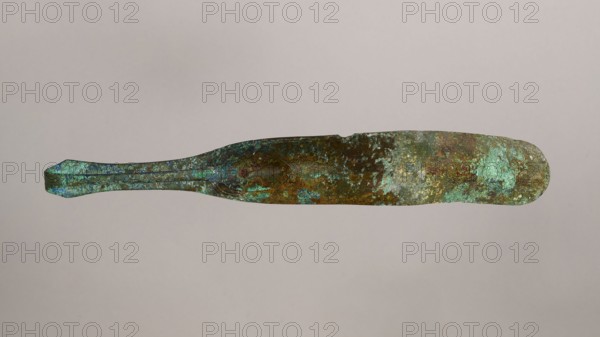 Scraper, Metal scraper with a typical, slightly curved shape. The item consists of a handle and a blade with a slightly rounded end. 6th–4th century BC, possibly Rome, tool; height 4.5 cm, width 28 cm, depth 11 cm, weight 0.11 kg., A long, narrow metal object with a flattened blade-like section and a tapered handle, showing a heavily weathered surface with patches of green and turquoise patina alongside areas of brown and dark rust-colored corrosion, set against a plain light beige-gray background.