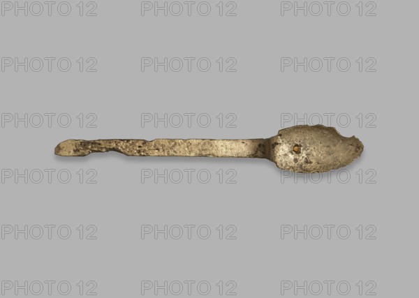 Medical tool fragment of strigil?, tool, height 12.8 cm, A single worn metal spoon lies horizontally against a plain light gray background; the spoon has a long, irregularly textured handle and an oval bowl with rough, pitted surfaces and an uneven edge, showing a tarnished metallic appearance in shades of dull gold, bronze, brown, and darker grayish spots.