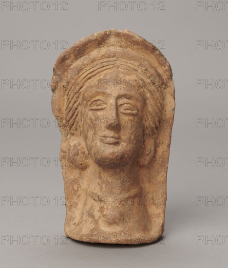 Votive head, 3rd–2nd century BC, Central Italy; sculpture; height 14.5 cm, width 9 cm, depth 8.5 cm, weight 0.5 kg; authenticity uncertain., A weathered terracotta head-shaped relief with a rounded top and a flat base, showing a stylized human face with closed or narrow eyes, a straight nose, and a small mouth, framed by textured hair or a headdress that forms layered ridges around the forehead and sides; the surface is rough and pitted with uneven edges and areas of wear, and the object rests against a plain background. Colors visible include various shades of brown, tan, beige, and hints of darker brown speckling.