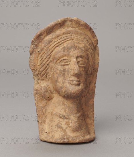 Votive head, 3rd–2nd century BC, Central Italy; sculpture; height 14.5 cm, width 9 cm, depth 8.5 cm, weight 0.5 kg; authenticity uncertain., A weathered terracotta sculpture head with a rounded top and stylized hair, displayed against a neutral gray background; the surface shows a rough texture with visible erosion and small pits, and the colors visible include shades of tan, light brown, reddish-brown, and darker brown speckling; the facial features are simplified with almond-shaped eyes, a pronounced nose, and full lips, and there is a suggestion of hair or a headdress framing the face and falling to the sides, with a short neck and a flattened back.