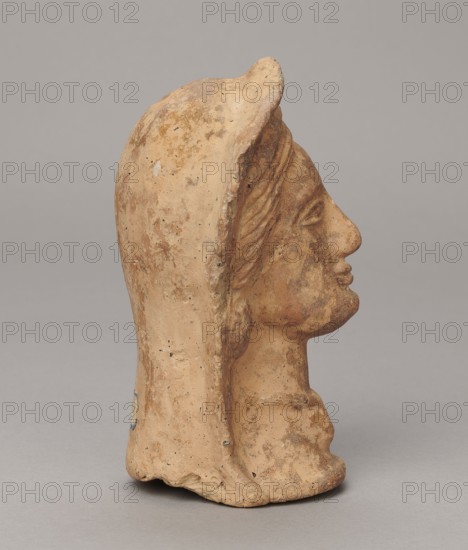 Votive head, 3rd–2nd century BC, Central Italy; sculpture; height 14.5 cm, width 9 cm, depth 8.5 cm, weight 0.5 kg; authenticity uncertain., The image shows a side view of a sculpted head made of a tan-brown material with weathered, rough surface texture and darker brown patina in creases and recessed areas; the head wears a hood that drapes down the back, and the facial features include a pronounced nose, closed lips, and a defined brow and cheek, all showing similar tan and brown coloration with occasional small dark specks; the background is a neutral light gray.