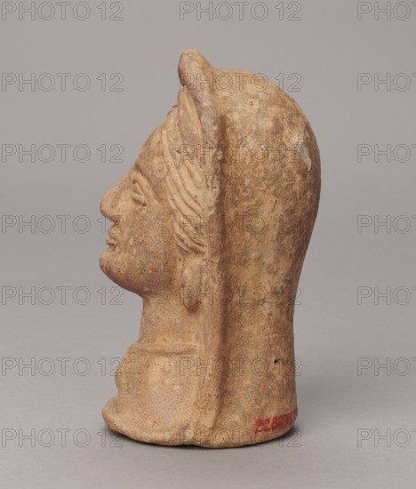Votive head, Madeyski, Antoni 1862-1939, 3rd–2nd century BC, Central Italy, sculpture; height 14.5 cm, width 9 cm, depth 8.5 cm, weight 0.5 kg, authenticity uncertain., A three-dimensional terracotta head viewed in profile with a few worn sculpted facial features including a nose, lips, chin, and a closed eye, showing a textured surface and areas of erosion. The head wears a molded headdress that extends down the back and sides, with a carved band or fold visible along the temple and a suggestion of stylized hair or feather-like ridges near the ear. The base is irregular and sits on a flat gray surface. The object displays a range of brown and tan tones with speckled darker brown and small pale spots, and there is a small area of red pigment near the base. The background is a uniform light gray.