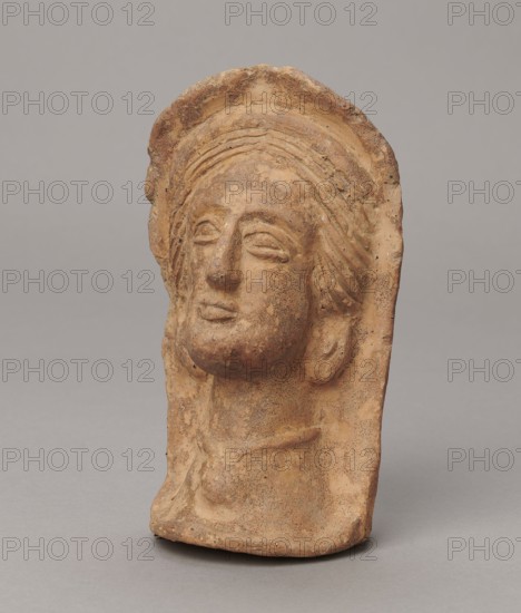 Votive head, 3rd–2nd century BC, Central Italy; sculpture; height 14.5 cm, width 9 cm, depth 8.5 cm, weight 0.5 kg; authenticity uncertain., A weathered terracotta head relief displayed against a plain gray background showing a sculpted face with closed eyes, a straight nose, and slightly parted lips; the hair is rendered in stylized waves framed by a rounded headpiece that extends down the sides, and the neck and upper chest are visible with a suggestion of a garment or collar. The surface shows a mottled texture with areas of roughness and small pits. The visible colors are earthy brown, tan, and hints of darker brown speckling, set against a neutral gray background.