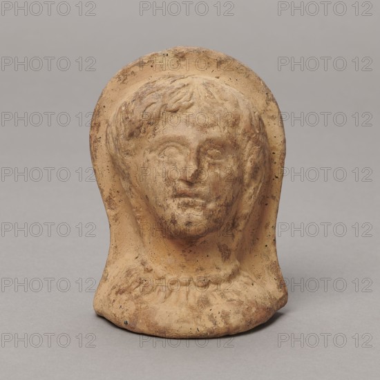Ex voto; maker, Hellenistic period, 3rd–2nd century BC circa 332–30 BC; possibly from Southern Etruria or Latium; bas-relief; height 9.5 cm, width 7 cm, depth 6.3 cm; weight 0.16 kg., A small sculpted head in a warm tan clay tone sits against a plain light gray background, showing a rounded face with short textured hair and a subtle halo-like rim around the top and sides; the surface shows darker brown weathered marks and small black speckles, with gently worn facial features and a simple rounded base that flares slightly at the bottom.