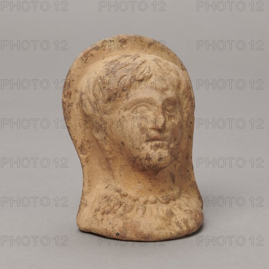 Ex voto, Antoni Madeyski 1862–1939 noted; Hellenistic period, 3rd–1st century BC 30 BC; Southern Etruria, / Latium ; bas-relief; height 9.5 cm, width 7 cm, depth 6.3 cm, weight 0.16 kg., A small sculpted head of a person made of a light brownish-tan material with darker brown mottling and spots, showing a rounded halo-like shape behind the head and stylized hair on the forehead, with subtle facial features including eyes, nose, and lips, and a suggestion of a necklace or decorative collar at the base; the object sits on a flat surface against a neutral gray background, and the visible colors include tan, brown, beige, and gray.