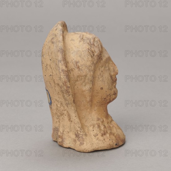 Ex voto, Madeyski, Antoni 1862–1939 - 30 BC; Hellenistic period; Southern Etruria, Latium ; bas-relief; height 9.5 cm, width 7 cm, depth 6.3 cm; weight 0.16 kg, A sculpted head in a beige to light tan clay color is shown in profile against a neutral gray background, with visible rough texture and patches of darker brown discoloration, and a small area of blue on the rear side; the face has a defined nose and chin, the head is draped with a raised hood or veil, and the base appears irregular and worn.