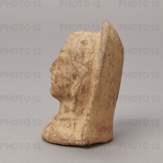 Ex voto, bas-relief, Hellenistic period, 332–30 BC; Southern Etruria, Latium ; height 9.5 cm, width 7 cm, depth 6.3 cm, weight 0.16 kg., A small sculpted profile of a human head and neck carved from a beige to light tan material with areas of darker brown patina and speckling, seen from the left side against a neutral gray background; the figure wears a rounded headdress or back piece that rises behind the head, and the surface shows textured tool marks and weathered roughness with subtle shadows and highlights.