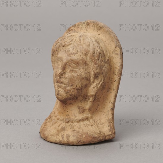 Ex voto, unknown creator; Madeyski, Antoni 1862–1939; 332; 30 BC; Hellenistic period; Southern Etruria, Latium ; bas-relief; height 9.5 cm; width 7 cm; depth 6.3 cm; weight 0.16 kg, A small sculpted head of a person made of a tan-colored material with darker brown patches and speckles, showing short textured hair, a rounded face with a straight nose and closed lips, a visible ear and a draped back element that curves around the head, and a base that is uneven and worn; colors visible include tan, brown, darker brown speckles, and a neutral gray background.
