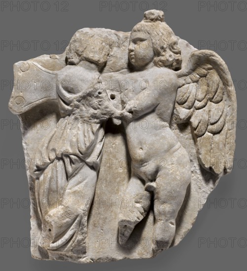 Fragment of a sarcophagus with a depiction of Eros and Psyche, unknown Roman workshop, Eros mythology, Psyche mythology, Roman sculpture, end of 2nd–beginning of 3rd century formerly dated 3rd century AD, Roman Empire 27 BC–476, sarcophagus, bas-relief, height 53 cm, width 48 cm, depth 8 cm, weight 22.5 kg, A carved stone relief showing a winged childlike figure and another partially visible figure in close contact; the winged figure is nude with rounded limbs and detailed feathered wings extending behind, and the other figure appears draped in flowing cloth with visible folds; the stone surface is weathered with chips and small holes; the dominant colors are pale beige and light gray with subtle darker gray shadows and traces of brownish discoloration.