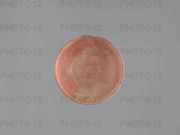 Plate, unknown Aswan workshop; flat plate on a low, ring-shaped foot. Rim poorly defined, rounded at the top with a vertical, slightly concave outer wall. Surfaces covered with red engobe and polished. Decoration executed by engraving and imprinting: three concentric grooves on the inner bottom, a roulette motif between the two outer grooves, and a central stamped cross in the form of a crux gemmata. Made of pink kaolin clay. 6th–7th century. Find location: Edfu. Vessel plate. Height 5.0–5.3 cm; diameter 26.8 cm rim, 13.7 cm bottom. Materials/technique: plates, Aswan pottery, Egyptian pottery, Egyptian sigillata, tableware, Coptic vessels, stamp impressions, stamped decoration, roulette decoration, kaolin clay., A round shallow dish with a worn terracotta surface sits against a smooth muted gray background; the dish shows circular ridges and areas of surface wear with a few lighter patches and subtle variations in reddish-brown and pale beige tones.