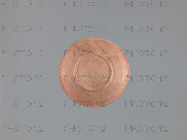 Plate, unknown Aswan workshop; flat plate on a low ring-shaped foot. Rim poorly defined, rounded at top, with a vertical, slightly concave outer wall. Surfaces covered with red engobe and polished; made of pink kaolin clay. Decoration by engraving and imprinting: three concentric grooves on inner bottom, roulette motif between outer grooves, central stamped cross in form of a crux gemmata. Egyptian pottery Egyptian sigillata, tableware/Coptic vessel; stamp impressions, stamped and roulette decoration. 6th–7th century; findspot Edfu. Vessel/plate; height 5.0–5.3 cm; diameter rim 26.8 cm; diameter bottom 13.7 cm., A round, shallow dish with a worn, matte surface in shades of terracotta and muted orange sits centered against a flat, uniform background of cool gray. The dish shows subtle variations in color with areas of lighter peach and darker rust, and faint circular ridges and scuff marks across its face. There are a few triangular repair lines and slightly darker patches near the rim,...