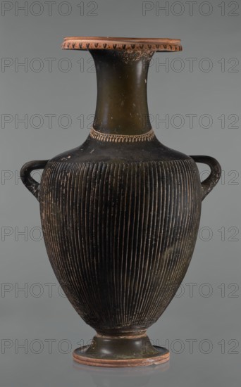 Black-finned hydria with fluting, unknown Apulian workshop. Hydria with fluting belly. Red clay. Black and shiny engobe firnis. The rim widens and turns outward, decorated with a painted ornament called an ovoid oxeye. The neck is long, cylindrical, decorated with a gilded garland painted at the base. The shoulders are clearly defined. The belly is oval, tapering towards the base, covered with fluting, impressed from the mold. The handles are cylindrical, attached vertically at shoulder height. Wide base. Preservation condition: cracked engobe in places, gilding worn away. 320–310 BC, Apulia, hydria vessel. Height 57 cm, width 24.7 cm., A tall ceramic vase with a narrow flared neck and a wide rounded body, featuring a pair of horizontal loop handles near the shoulder; the vase surface shows a ribbed texture on the body and a smoother neck and base with small areas of wear and speckling. The colors visible include deep black on most of the body and neck, earthy terracotta on the rim and...