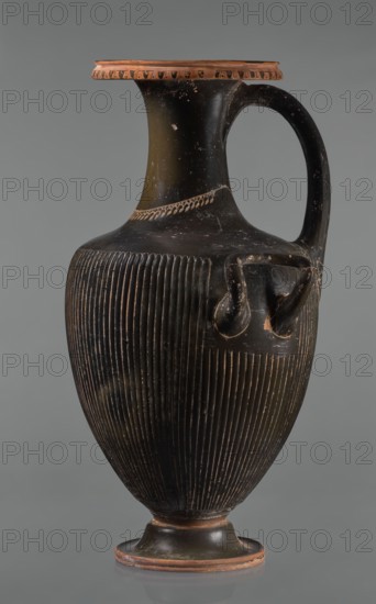 Black-finned hydria with fluting, unknown Apulian workshop. Hydria with fluting belly. Red clay. Black and shiny engobe firnis. The rim widens and turns outward, decorated with a painted ornament called an ovoid oxeye. The neck is long, cylindrical, decorated with a gilded garland painted at the base. The shoulders are clearly defined. The belly is oval, tapering towards the base, covered with fluting, impressed from the mold. The handles are cylindrical, attached vertically at shoulder height. Wide base. Preservation condition: cracked engobe in places, gilding worn away. 320–310 BC, Apulia, hydria vessel. Height 57 cm, width 24.7 cm., A tall, narrow ceramic jug with a rounded body and a single curved handle is shown against a plain grey background; the jug has a flared rim and sits on a small circular foot, and its surface is primarily black with visible areas of worn reddish-brown along the rim, foot, and some scuffed spots; the body is decorated with a pattern of vertical incised g...