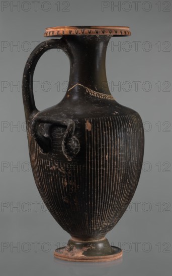 Black-finned hydria with fluting, unknown Apulian workshop. Hydria with fluting belly. Red clay. Black and shiny engobe firnis. The rim widens and turns outward, decorated with a painted ornament called an ovoid oxeye. The neck is long, cylindrical, decorated with a gilded garland painted at the base. The shoulders are clearly defined. The belly is oval, tapering towards the base, covered with fluting, impressed from the mold. The handles are cylindrical, attached vertically at shoulder height. Wide base. Preservation condition: cracked engobe in places, gilding worn away. 320–310 BC, Apulia, hydria, vessel; height 57 cm, width 24.7 cm., A tall, narrow-handled ceramic vessel with a dark black surface and areas of worn terracotta color, featuring a rounded body that tapers to a small foot, a flared rim with a band of lighter terracotta around the edge, a curved single handle from rim to shoulder, a pair of small looped side handles on the shoulder, vertical incised lines covering the lo...