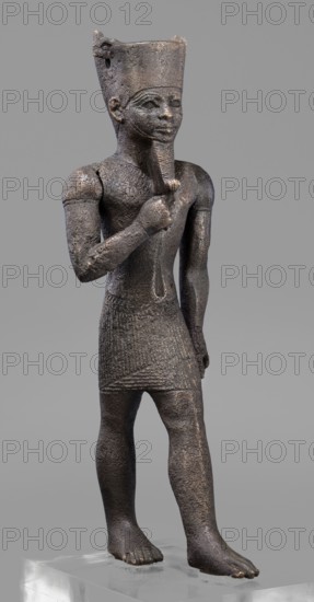 Figurine of the god Amun, Amun mythology, Egyptian deities, male figurines, Egyptian art, Once holding a khepesh, ca. 1069–ca. 747 BC; Third Intermediate Period, Egypt, sculpture, height 19.6 cm, width 6.2 cm, A dark brown textured statue of a standing human figure on a clear rectangular base, wearing a tall flat-topped headdress and a short patterned skirt, with one arm bent so the hand touches a stylized beard and the other arm hanging by the side, the face showing almond-shaped eyes and a calm expression, the surface showing rough, worn areas and lighter brown highlights against a neutral gray background.